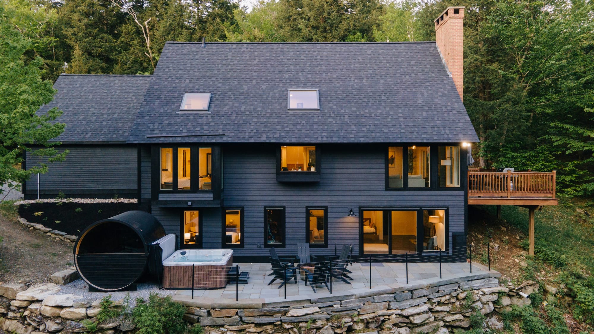 A Far & Away managed vacation rental near Stratton Mountain at dusk, with a barrel sauna, hot tub and lit windows against the Vermont woods